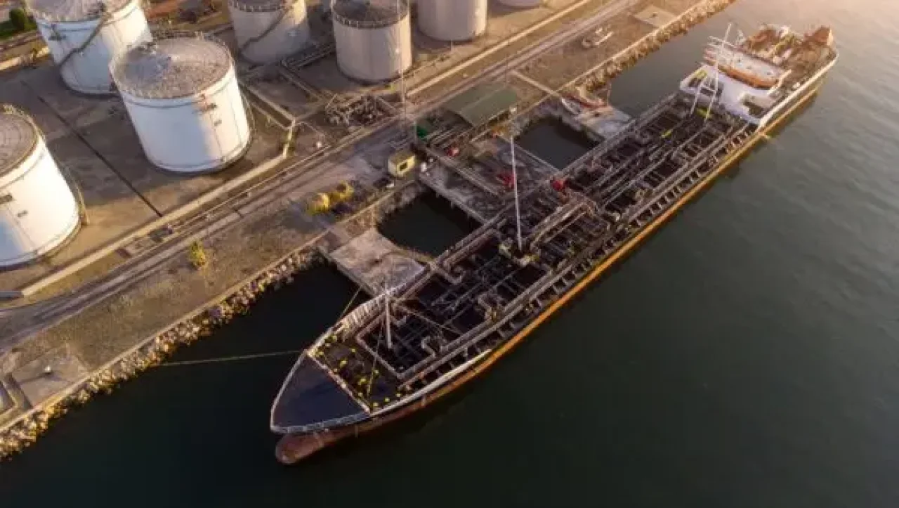 An aerial view showcases a bustling port scene, featuring a cargo ship docked near white storage tanks, with a golden sunset casting long shadows.