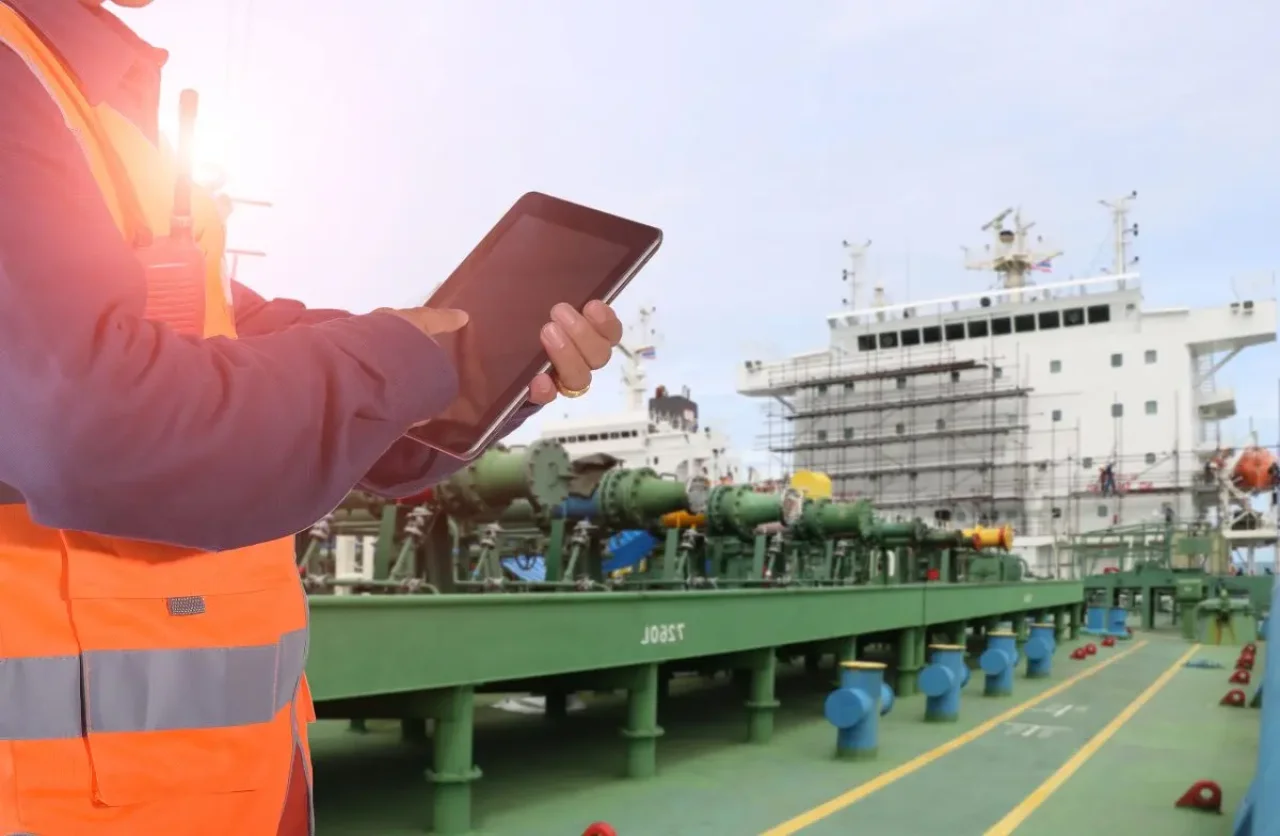 A worker in an orange vest uses a tablet on a green platform attached to a white ship with blue and yellow accents, while blue and red cones indicate ongoing work.