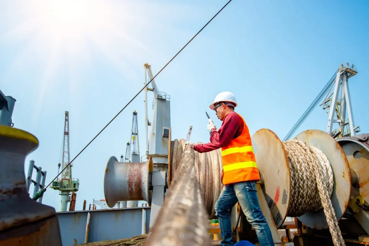 A construction worker, dressed in safety gear, communicates on a rope connected to a crane on a bustling construction site under a clear blue sky.