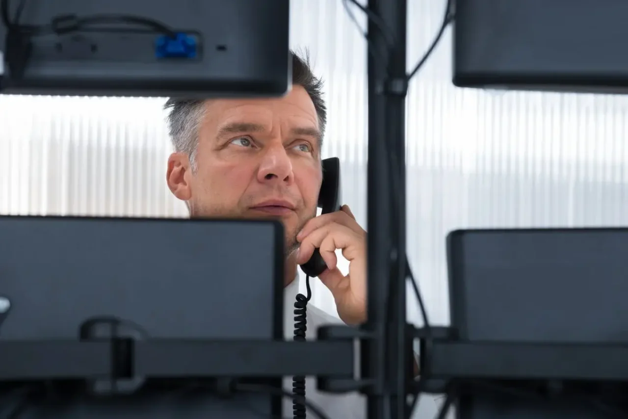 A man with gray hair, dressed professionally, converses on a black phone between two black computer monitors, framed by white curtains.