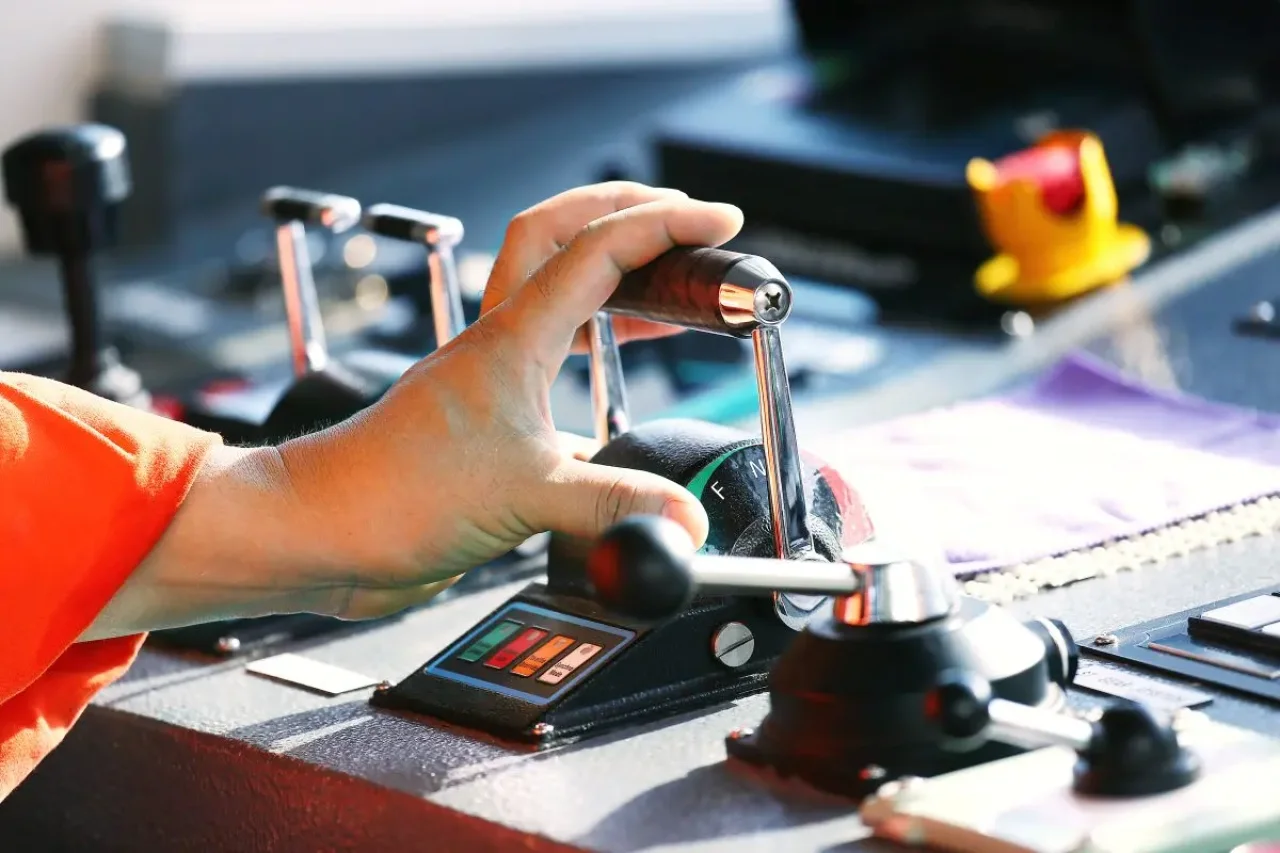 A hand, wearing a red shirt, grips a black lever with a silver handle, controlling a machinery rig with a purple and black control panel.