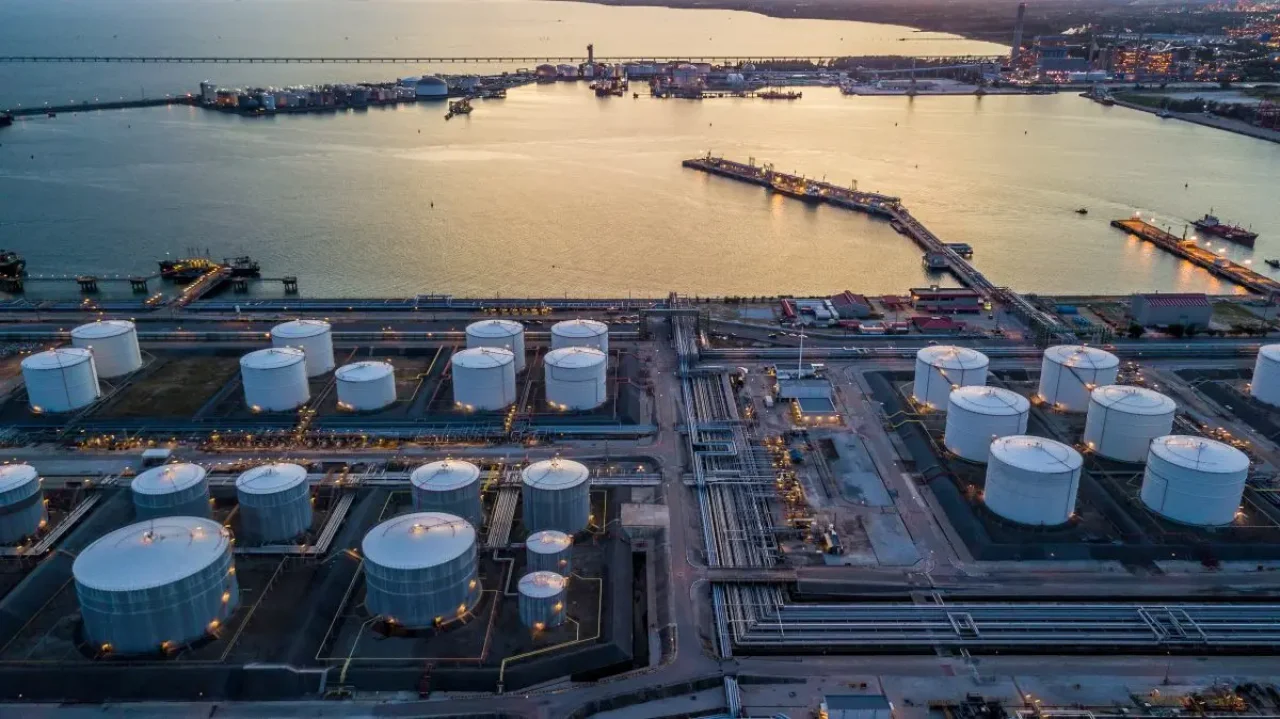 An aerial view of an oil refinery at sunset showcases large white tanks, intricate pipes, and buildings, with a calm water and city skyline in the background.